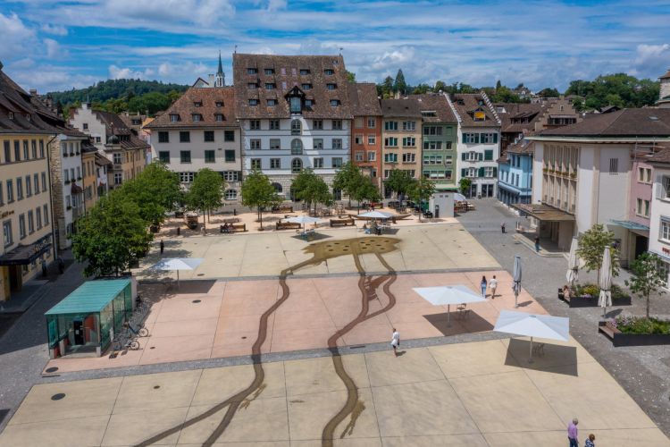 Luftaufnahme des Herrenackerplatzes in Schaffhausen an einem sonnigen Tag mit umliegenden Häusern. Viele Sitzgelegenheiten, das Wasserspiel und Bäume sind zu sehen.